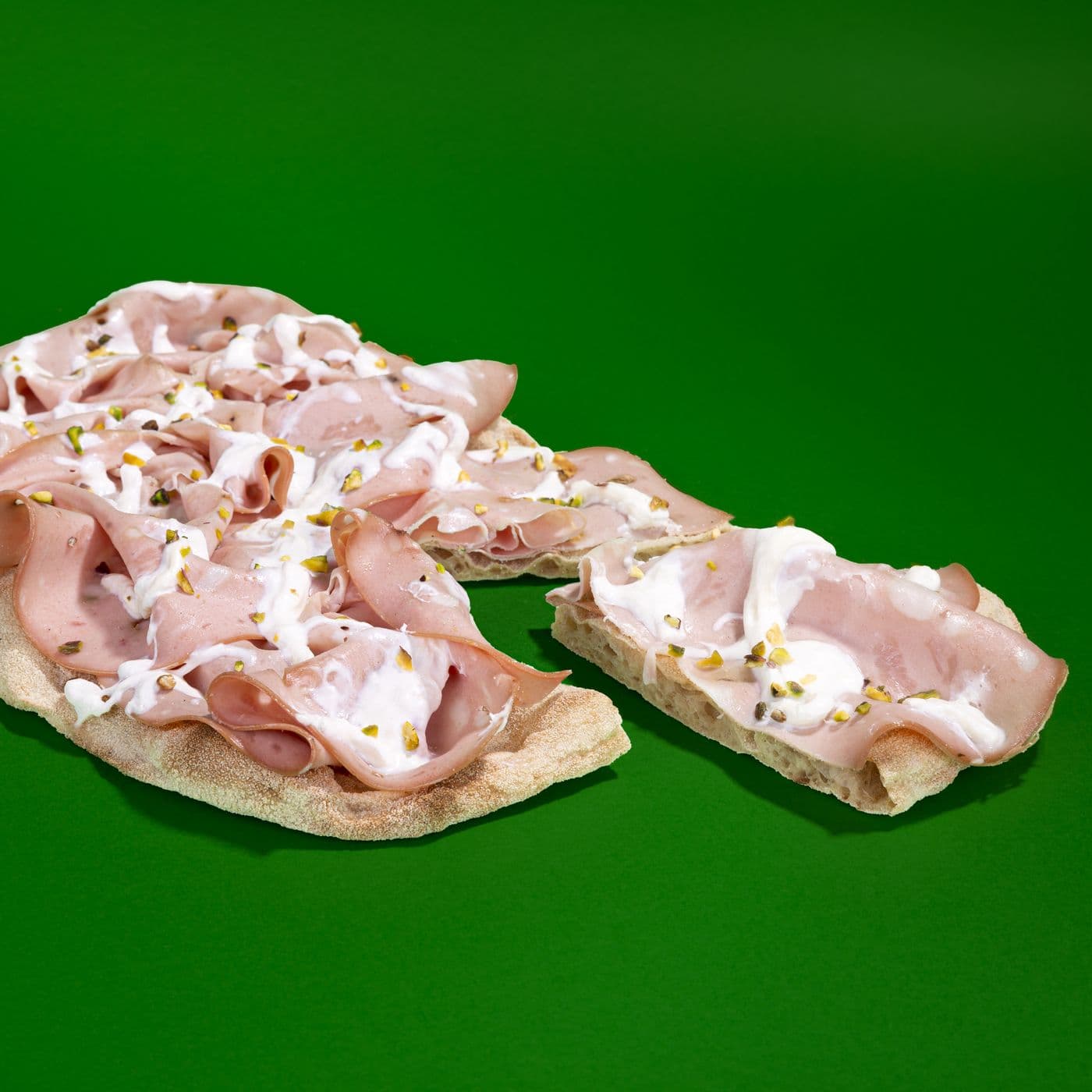 Pinsa con mortadella, stracciatella e pistacchi divisa a metà, smezzata su fondo verde